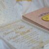 A high-angle shot featuring an open peach-colored wedding invitation box set on a white surface with draped white fabric and a gold lace cuff. On the left, the closed lid of the box displays a central gold plaque with the monogram "R & P," framed by purple scrollwork accented with pearls. On the right, the open box base reveals a formal acrylic invitation for "James Parker & Karen Brett" printed with gold text. A gold RSVP card with matching scrollwork is partially tucked into a pocket in the open lid.