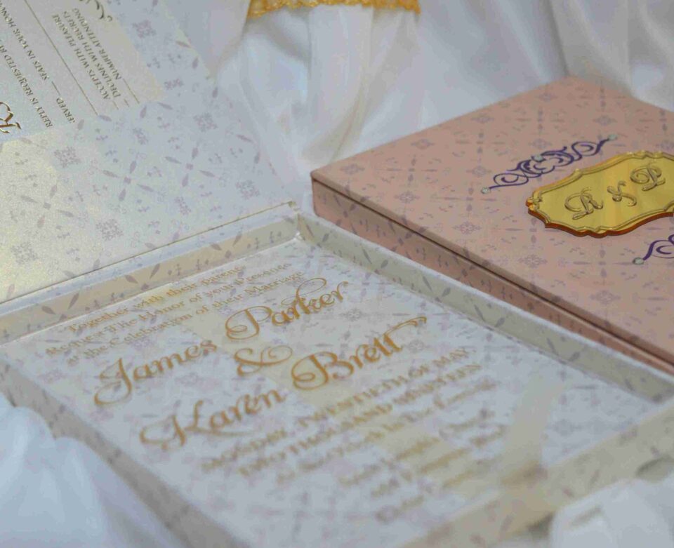 A high-angle shot featuring an open peach-colored wedding invitation box set on a white surface with draped white fabric and a gold lace cuff. On the left, the closed lid of the box displays a central gold plaque with the monogram "R & P," framed by purple scrollwork accented with pearls. On the right, the open box base reveals a formal acrylic invitation for "James Parker & Karen Brett" printed with gold text. A gold RSVP card with matching scrollwork is partially tucked into a pocket in the open lid.