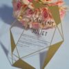 A transparent acrylic wedding invitation with a striking gold geometric frame is displayed leaning against a bouquet of soft peach and pink roses. The frame features sharp, crystalline lines and solid gold triangular accents in the corners.