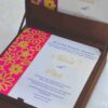 An open, chocolate-brown fabric invitation box displaying a formal wedding reception card. The card features a vibrant magenta left border with a gold laser-cut floral pattern. The main white section of the card includes elegant gold and purple typography for the names "Aakash with Preeti" and details for an event held in Hyderabad. The lid of the box is propped open, showing a matching design inside.