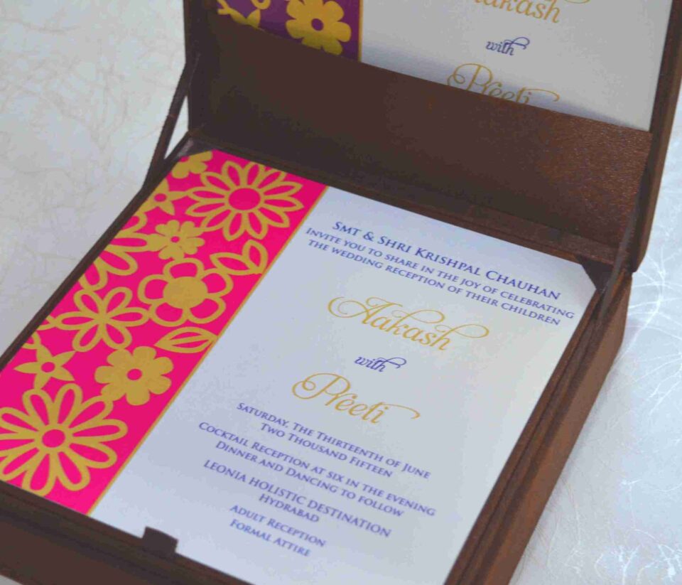 An open, chocolate-brown fabric invitation box displaying a formal wedding reception card. The card features a vibrant magenta left border with a gold laser-cut floral pattern. The main white section of the card includes elegant gold and purple typography for the names "Aakash with Preeti" and details for an event held in Hyderabad. The lid of the box is propped open, showing a matching design inside.