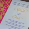 An open, chocolate-brown fabric invitation box displaying a formal wedding reception card. The card features a vibrant magenta left border with a gold laser-cut floral pattern. The main white section of the card includes elegant gold and purple typography for the names "Aakash with Preeti" and details for an event held in Hyderabad. The lid of the box is propped open, showing a matching design inside.