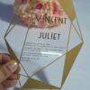 A transparent acrylic wedding invitation with a striking gold geometric frame is displayed leaning against a bouquet of soft peach and pink roses. The frame features sharp, crystalline lines and solid gold triangular accents in the corners.