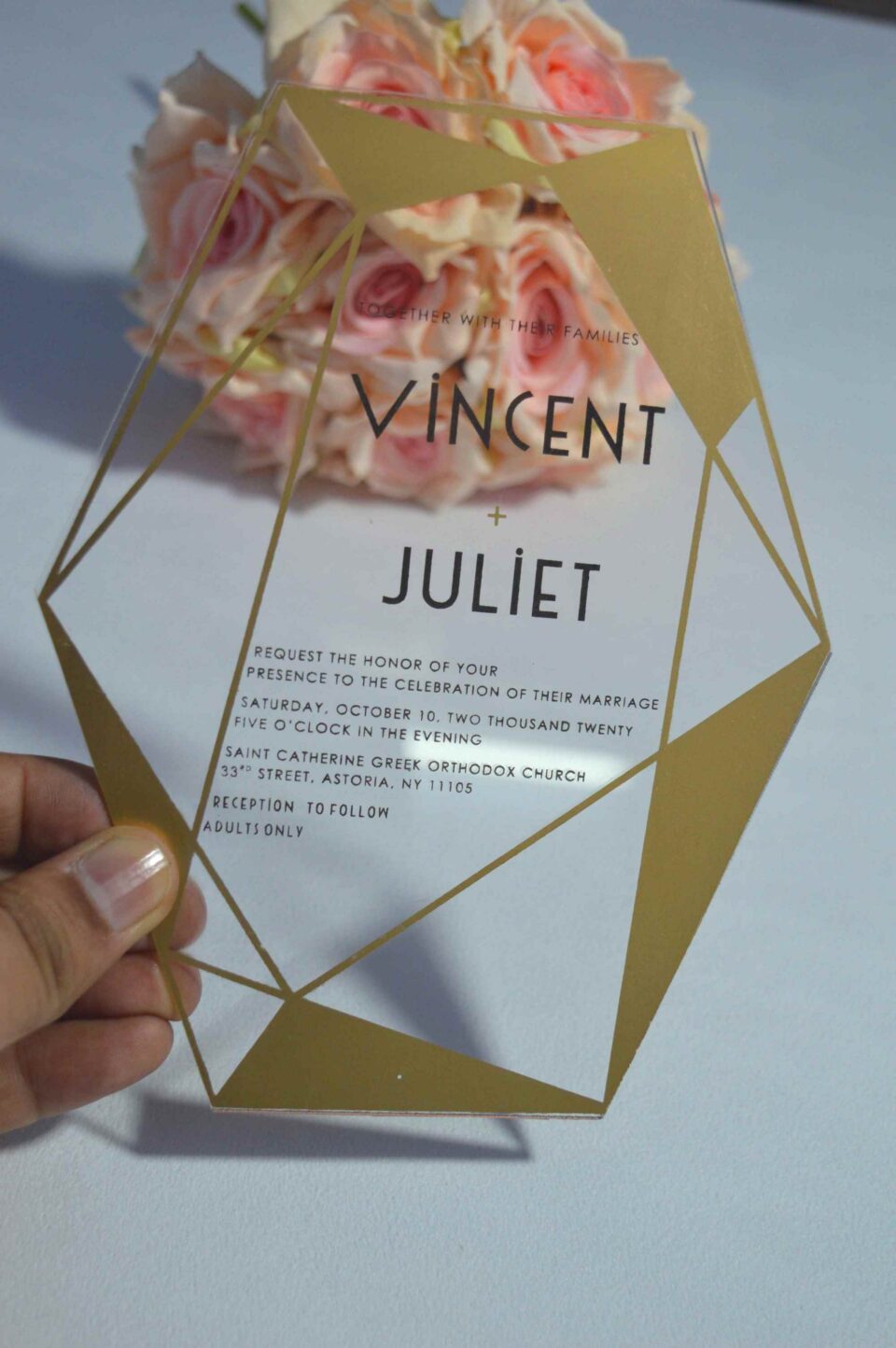 A transparent acrylic wedding invitation with a striking gold geometric frame is displayed leaning against a bouquet of soft peach and pink roses. The frame features sharp, crystalline lines and solid gold triangular accents in the corners.