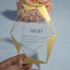 A transparent acrylic wedding invitation with a striking gold geometric frame is displayed leaning against a bouquet of soft peach and pink roses. The frame features sharp, crystalline lines and solid gold triangular accents in the corners.