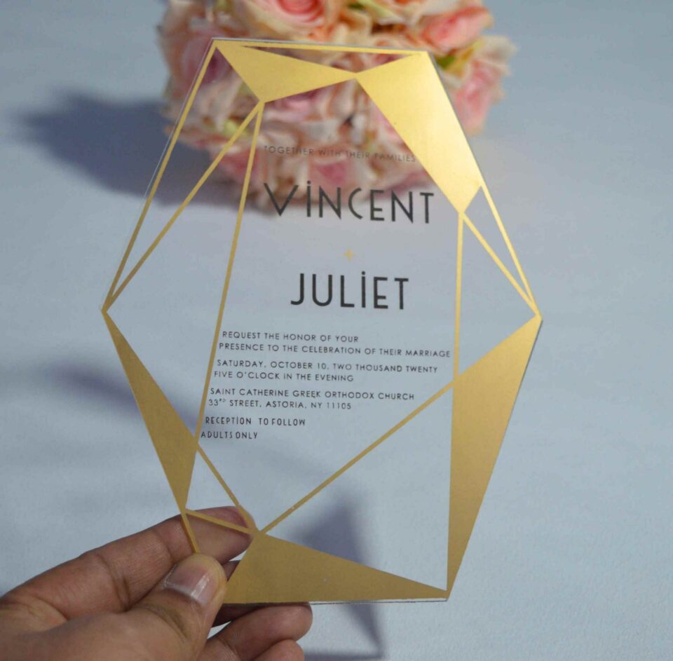 A transparent acrylic wedding invitation with a striking gold geometric frame is displayed leaning against a bouquet of soft peach and pink roses. The frame features sharp, crystalline lines and solid gold triangular accents in the corners.