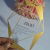 A transparent acrylic wedding invitation with a striking gold geometric frame is displayed leaning against a bouquet of soft peach and pink roses. The frame features sharp, crystalline lines and solid gold triangular accents in the corners.