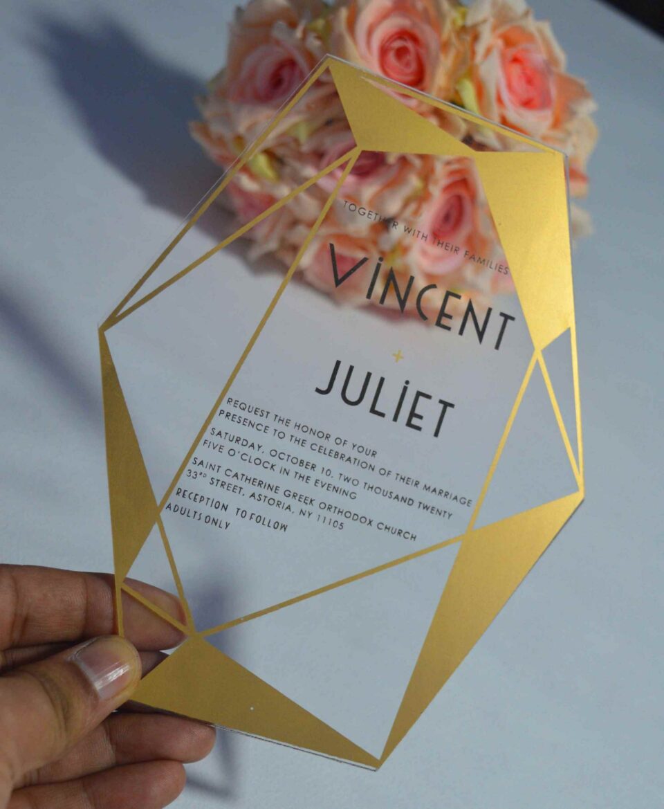 A transparent acrylic wedding invitation with a striking gold geometric frame is displayed leaning against a bouquet of soft peach and pink roses. The frame features sharp, crystalline lines and solid gold triangular accents in the corners.