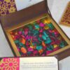 An open, chocolate-brown invitation box with a gold-lined interior filled with colorful potpourri in shades of magenta, teal, orange, and yellow. The lid is propped open, showing a glimpse of an invitation card for "Aakash with Preeti." In the foreground, a matching invitation insert is visible, and to the right, a section of a gold laser-cut decorative panel sits on the white textured surface.