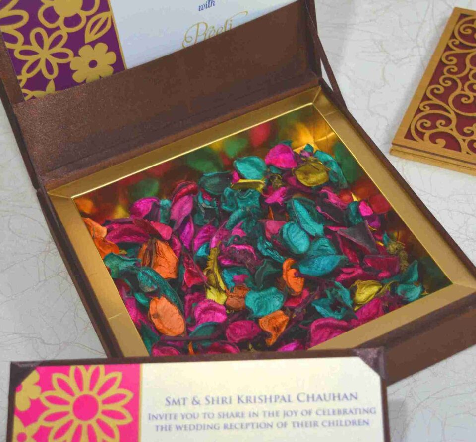 An open, chocolate-brown invitation box with a gold-lined interior filled with colorful potpourri in shades of magenta, teal, orange, and yellow. The lid is propped open, showing a glimpse of an invitation card for "Aakash with Preeti." In the foreground, a matching invitation insert is visible, and to the right, a section of a gold laser-cut decorative panel sits on the white textured surface.