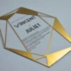 A clear acrylic wedding invitation in a sharp, geometric diamond shape lies flat on a light gray surface. The invitation features a striking border made of metallic gold triangles and intersecting lines that frame the central text.