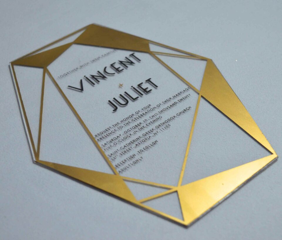 A clear acrylic wedding invitation in a sharp, geometric diamond shape lies flat on a light gray surface. The invitation features a striking border made of metallic gold triangles and intersecting lines that frame the central text.