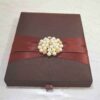 A high-angle shot of a rectangular, dark brown silk invitation box resting on a light-colored marbled surface. A wide, burgundy satin ribbon is wrapped horizontally around the center, gathered in the middle by a large, circular brooch. The brooch features a cluster of creamy white pearls interspersed with small, gold-toned floral accents set with tiny crystals. A small ribbon pull-tab is visible at the bottom edge of the box.