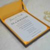 An angled, close-up shot of a wedding invitation suite. A brown silk box with a vibrant yellow-orange satin lining holds a white invitation card that reads "James & Wallis ARE GETTING MARRIED!" in elegant black typography. Below the box, an RSVP card and a detail card with gold-foil lettering for "Victoria and Patric" are displayed. To the left, a string of large, creamy white pearls adds a touch of luxury, all resting on a light-colored marbled surface.