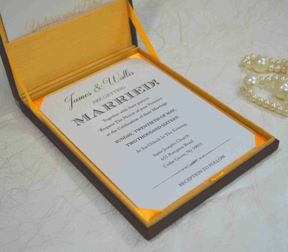 An angled, close-up shot of a wedding invitation suite. A brown silk box with a vibrant yellow-orange satin lining holds a white invitation card that reads "James & Wallis ARE GETTING MARRIED!" in elegant black typography. Below the box, an RSVP card and a detail card with gold-foil lettering for "Victoria and Patric" are displayed. To the left, a string of large, creamy white pearls adds a touch of luxury, all resting on a light-colored marbled surface.