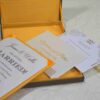 An angled, close-up shot of a wedding invitation suite. A brown silk box with a vibrant yellow-orange satin lining holds a white invitation card that reads "James & Wallis ARE GETTING MARRIED!" in elegant black typography. Below the box, an RSVP card and a detail card with gold-foil lettering for "Victoria and Patric" are displayed. To the left, a string of large, creamy white pearls adds a touch of luxury, all resting on a light-colored marbled surface.
