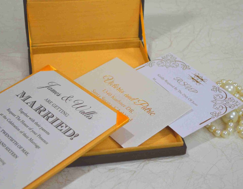 An angled, close-up shot of a wedding invitation suite. A brown silk box with a vibrant yellow-orange satin lining holds a white invitation card that reads "James & Wallis ARE GETTING MARRIED!" in elegant black typography. Below the box, an RSVP card and a detail card with gold-foil lettering for "Victoria and Patric" are displayed. To the left, a string of large, creamy white pearls adds a touch of luxury, all resting on a light-colored marbled surface.