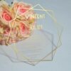Transparent acrylic wedding invitation with a sleek, rectangular shape is shown leaning against a lush bouquet of light pink and peach roses. The invitation features a modern gold geometric border made of thin, intersecting lines and solid gold accents at the bottom. The text is printed in gold.