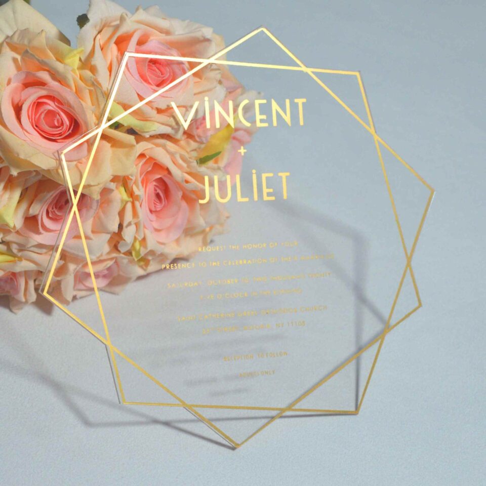 Transparent acrylic wedding invitation with a sleek, rectangular shape is shown leaning against a lush bouquet of light pink and peach roses. The invitation features a modern gold geometric border made of thin, intersecting lines and solid gold accents at the bottom. The text is printed in gold.