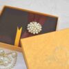 A high-angle shot of a rectangular, dark brown silk invitation box resting on a light-colored marbled surface. A wide, burgundy satin ribbon is wrapped horizontally around the center, gathered in the middle by a large, circular brooch. The brooch features a cluster of creamy white pearls interspersed with small, gold-toned floral accents set with tiny crystals. A small ribbon pull-tab is visible at the bottom edge of the box.