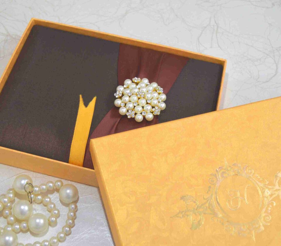 A high-angle shot of a rectangular, dark brown silk invitation box resting on a light-colored marbled surface. A wide, burgundy satin ribbon is wrapped horizontally around the center, gathered in the middle by a large, circular brooch. The brooch features a cluster of creamy white pearls interspersed with small, gold-toned floral accents set with tiny crystals. A small ribbon pull-tab is visible at the bottom edge of the box.