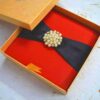 A square, vibrant red silk-textured invitation box shown from a slightly elevated angle. A wide black satin ribbon wraps horizontally across the center of the box, gathered at the middle by a large, circular gold-toned brooch composed of numerous cream pearls and small sparkling crystals. The box sits on a white, textured surface scattered with a few dried yellow flower petals. The bright lighting emphasizes the rich color of the fabric and the luster of the pearls.
