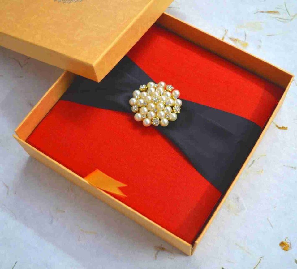 A square, vibrant red silk-textured invitation box shown from a slightly elevated angle. A wide black satin ribbon wraps horizontally across the center of the box, gathered at the middle by a large, circular gold-toned brooch composed of numerous cream pearls and small sparkling crystals. The box sits on a white, textured surface scattered with a few dried yellow flower petals. The bright lighting emphasizes the rich color of the fabric and the luster of the pearls.