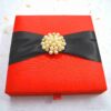 A square, vibrant red silk-textured invitation box shown from a slightly elevated angle. A wide black satin ribbon wraps horizontally across the center of the box, gathered at the middle by a large, circular gold-toned brooch composed of numerous cream pearls and small sparkling crystals. The box sits on a white, textured surface scattered with a few dried yellow flower petals. The bright lighting emphasizes the rich color of the fabric and the luster of the pearls.