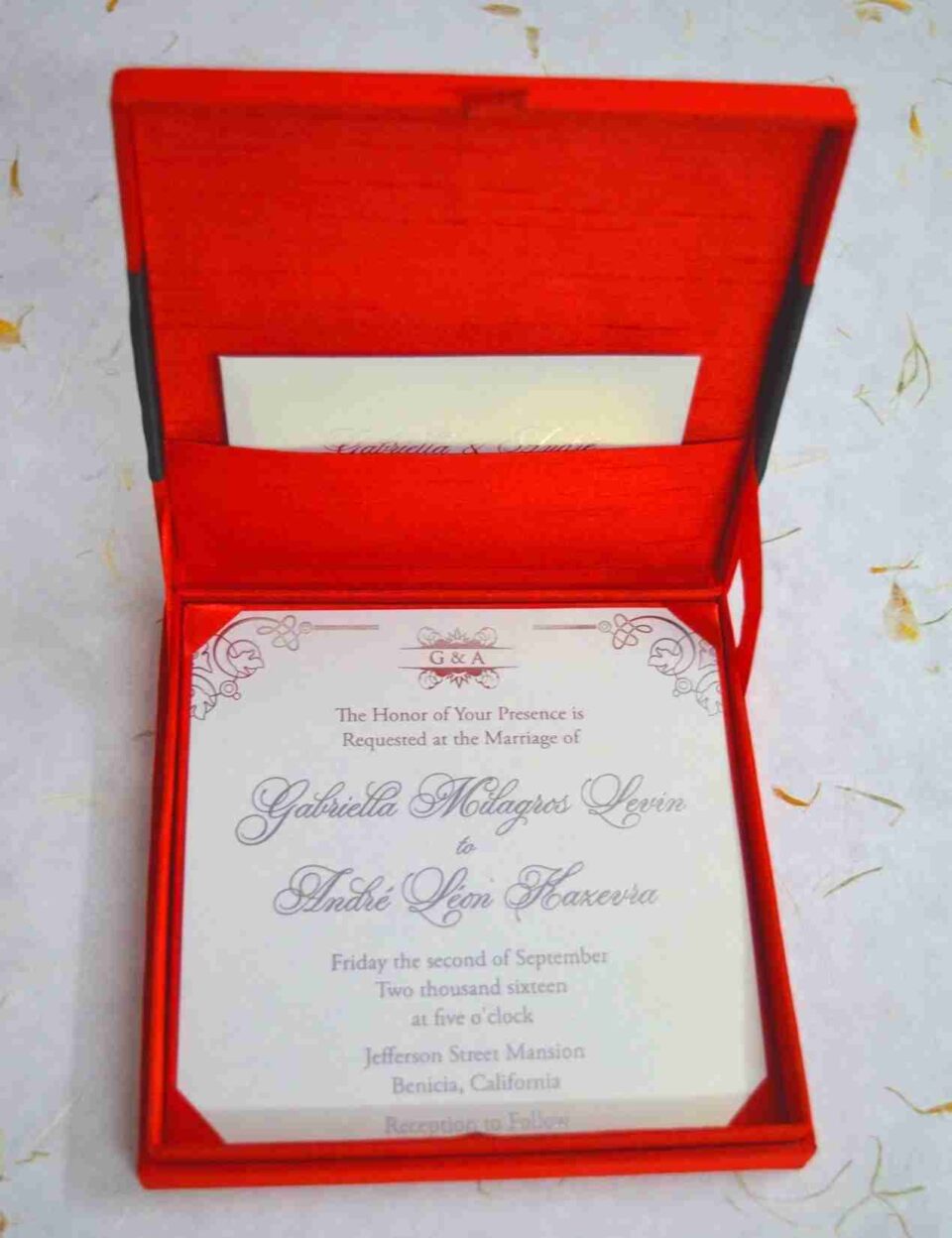 An open, square wedding invitation box in vibrant red silk. The main invitation card is nestled in the base, featuring the names "Gabriella Milagros Levin" and "André Leon Hazevra" in elegant grey script, topped with a small "G & A" monogram and decorative corner flourishes. The inner lid of the box contains a pocket holding a matching white card. The box is set on a white textured surface scattered with delicate yellow petals, creating a formal and colorful presentation.