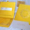 A high-angle shot of a luxurious wedding invitation suite in vibrant gold, arranged on a soft white fabric. * **On the left:** An open gold silk invitation box. The inner lid features a pocket holding a white RSVP card with gold floral accents. The bottom compartment contains a clear acrylic invitation card. * **On the right:** The closed lid of the gold box, featuring an embossed circular emblem with ornate scrollwork, the names "James Parker & Karen Brett," the date "09.09.2019," and small embedded crystals. * **In the foreground:** A white mailing envelope addressed to "Aleeya Rahman" in gold lettering. * **In the background:** Decorative gold lace pieces with small blue crystals are scattered for additional texture.