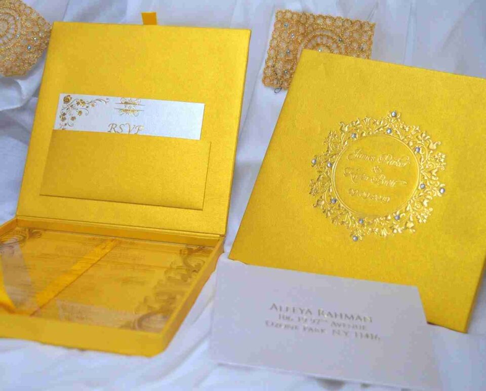 A high-angle shot of a luxurious wedding invitation suite in vibrant gold, arranged on a soft white fabric. * **On the left:** An open gold silk invitation box. The inner lid features a pocket holding a white RSVP card with gold floral accents. The bottom compartment contains a clear acrylic invitation card. * **On the right:** The closed lid of the gold box, featuring an embossed circular emblem with ornate scrollwork, the names "James Parker & Karen Brett," the date "09.09.2019," and small embedded crystals. * **In the foreground:** A white mailing envelope addressed to "Aleeya Rahman" in gold lettering. * **In the background:** Decorative gold lace pieces with small blue crystals are scattered for additional texture.
