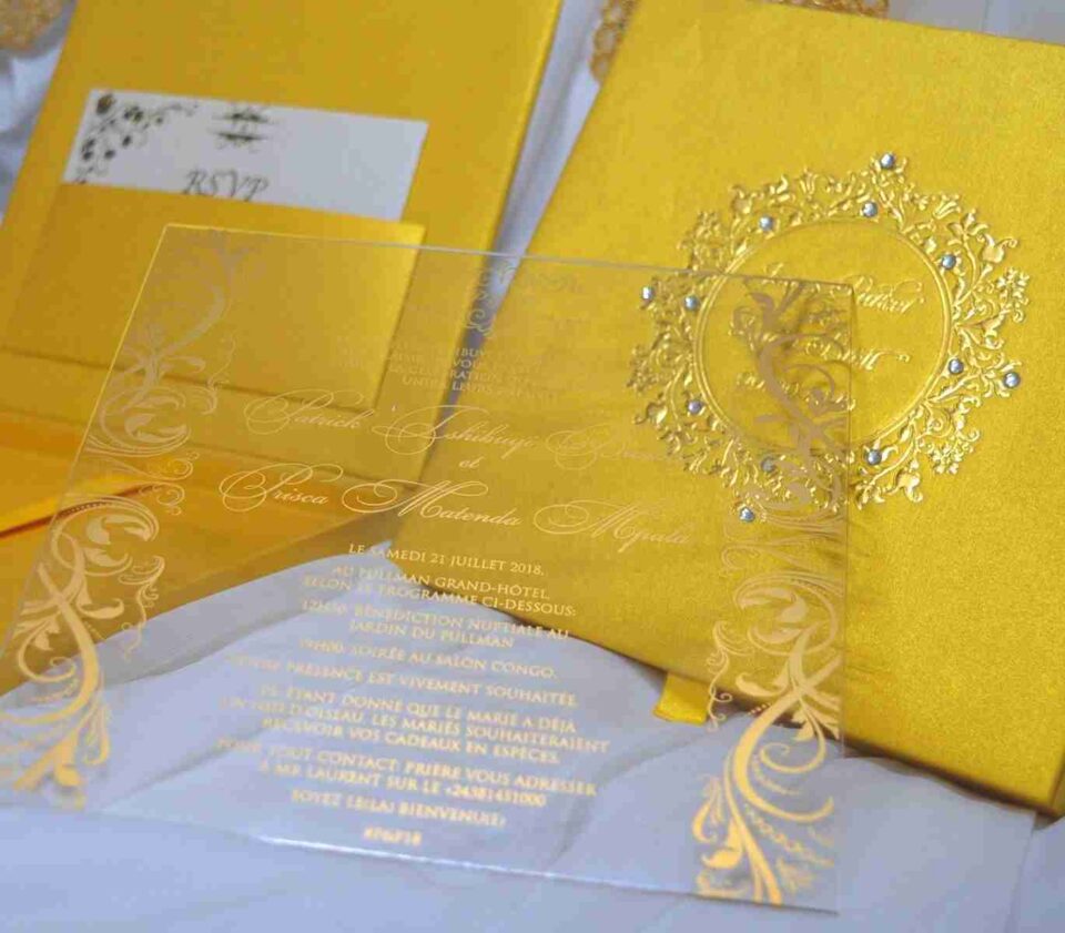 A high-angle, close-up shot of a luxury wedding invitation suite in shimmering gold. A clear acrylic invitation card with gold-foil script and ornate corner flourishes for "Patrick and Prisca" is leaned against its matching gold silk box. The lid of the box, visible behind the acrylic, features a debossed circular crest decorated with crystals. To the left, the open base of the box shows a yellow-gold satin lining and an RSVP card tucked into an interior pocket. The entire suite rests on a soft, white textured fabric.