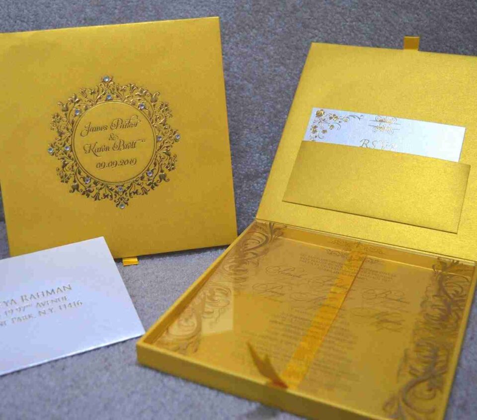 A high-angle shot of a luxurious wedding invitation suite in vibrant gold, arranged on a soft white fabric. * **On the left:** An open gold silk invitation box. The inner lid features a pocket holding a white RSVP card with gold floral accents. The bottom compartment contains a clear acrylic invitation card. * **On the right:** The closed lid of the gold box, featuring an embossed circular emblem with ornate scrollwork, the names "James Parker & Karen Brett," the date "09.09.2019," and small embedded crystals. * **In the foreground:** A white mailing envelope addressed to "Aleeya Rahman" in gold lettering. * **In the background:** Decorative gold lace pieces with small blue crystals are scattered for additional texture.