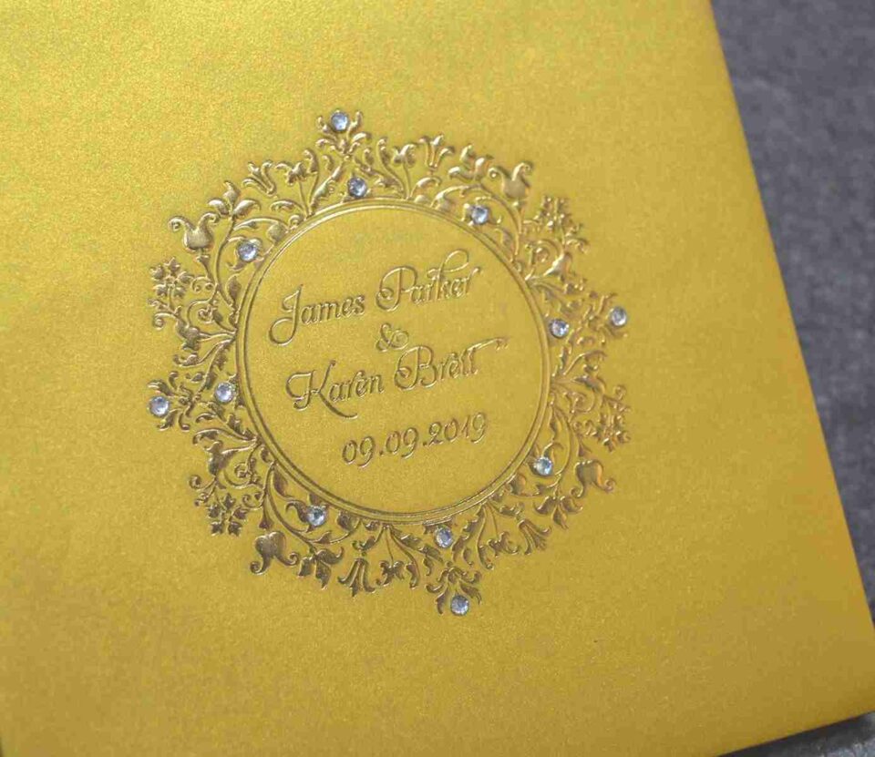 A close-up, angled shot of a shimmering gold invitation card. The center features a detailed, embossed circular emblem with ornate scrollwork and floral patterns. Inside the circle, the names "James Parker & Karen Brett" and the date "09.09.2019" are elegantly engraved. Small, sparkling crystals are delicately embedded within the surrounding gold flourishes, adding a touch of brilliance to the design.