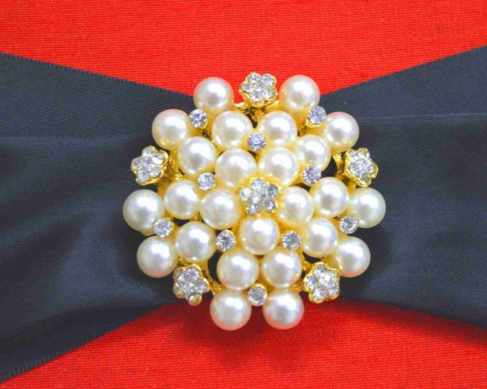 A square, vibrant red silk-textured invitation box shown from a slightly elevated angle. A wide black satin ribbon wraps horizontally across the center of the box, gathered at the middle by a large, circular gold-toned brooch composed of numerous cream pearls and small sparkling crystals. The box sits on a white, textured surface scattered with a few dried yellow flower petals. The bright lighting emphasizes the rich color of the fabric and the luster of the pearls.