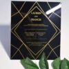 A square, elegant wedding invitation featuring a black acrylic or high-gloss finish with gold geometric line patterns and text. The invitation is held by a hand in the bottom-left corner against a plain white background, with green leaves resting over the .The visible in the soft-focus background on the left.