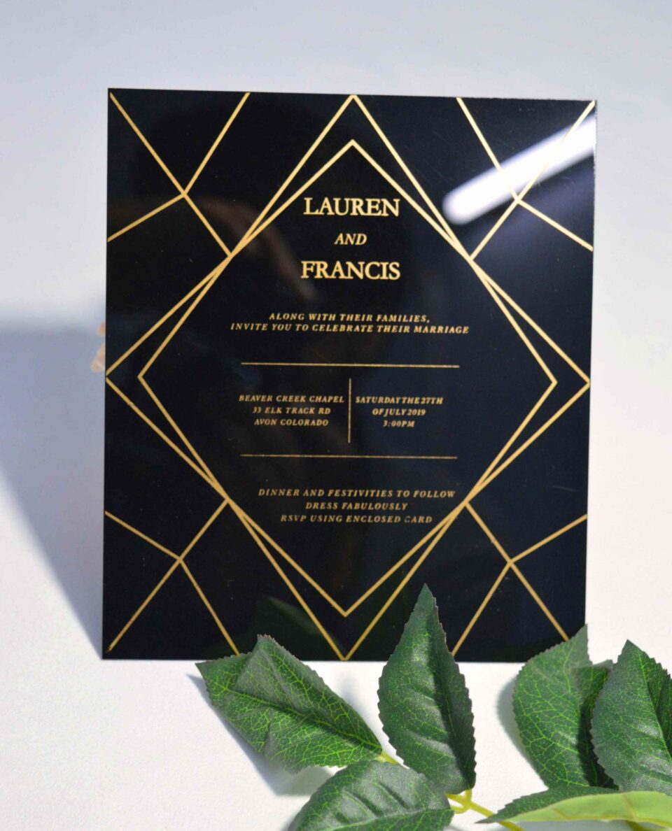A square, elegant wedding invitation featuring a black acrylic or high-gloss finish with gold geometric line patterns and text. The invitation is held by a hand in the bottom-left corner against a plain white background, with green leaves resting over the .The visible in the soft-focus background on the left.