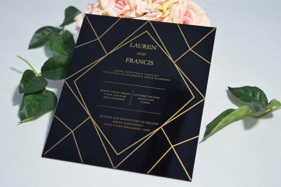 A square, elegant wedding invitation featuring a black acrylic or high-gloss finish with gold geometric line patterns and text. The invitation is held by a hand in the bottom-left corner against a plain white background, with green leaves resting over the .The visible in the soft-focus background on the left.