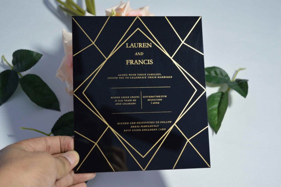 A square, elegant wedding invitation featuring a black acrylic or high-gloss finish with gold geometric line patterns and text. The invitation is held by a hand in the bottom-left corner against a plain white background, with a bouquet of light pink roses and A square, elegant wedding invitation featuring a black acrylic or high-gloss finish with gold geometric line patterns and text. The invitation is held by a hand in the bottom-left corner against a plain white background, with a bouquet of light pink roses and green leaves resting over the corner.