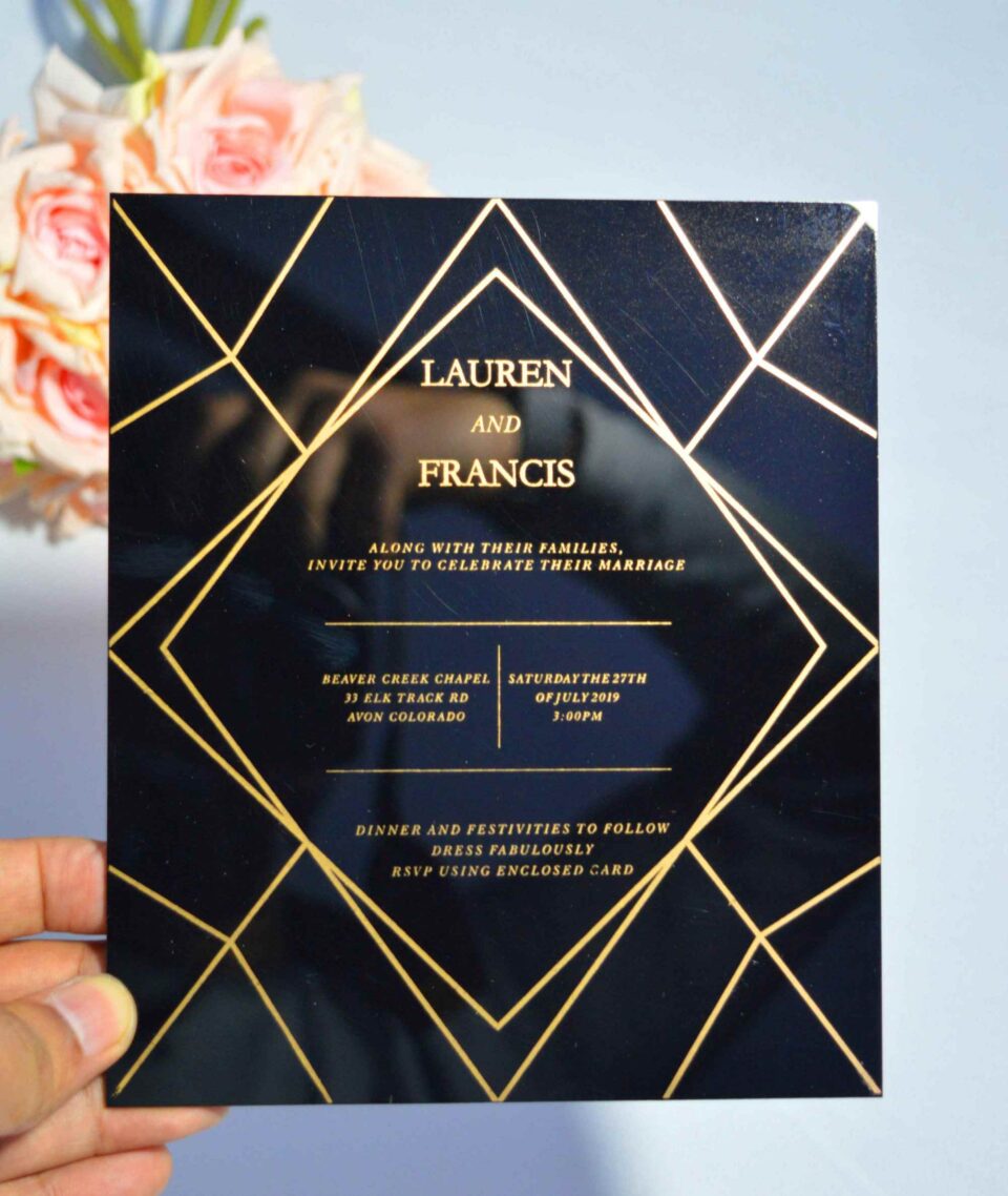 A square, elegant wedding invitation featuring a black acrylic or high-gloss finish with gold geometric line patterns and text. The invitation is held by a hand in the bottom-left corner against a plain white background, with a bouquet of light pink roses visible in the soft-focus background on the left.