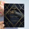 A square, elegant wedding invitation featuring a black acrylic or high-gloss finish with gold geometric line patterns and text. The invitation is held by a hand in the bottom-left corner against a plain white background, with a bouquet of light pink roses visible in the soft-focus background on the left.