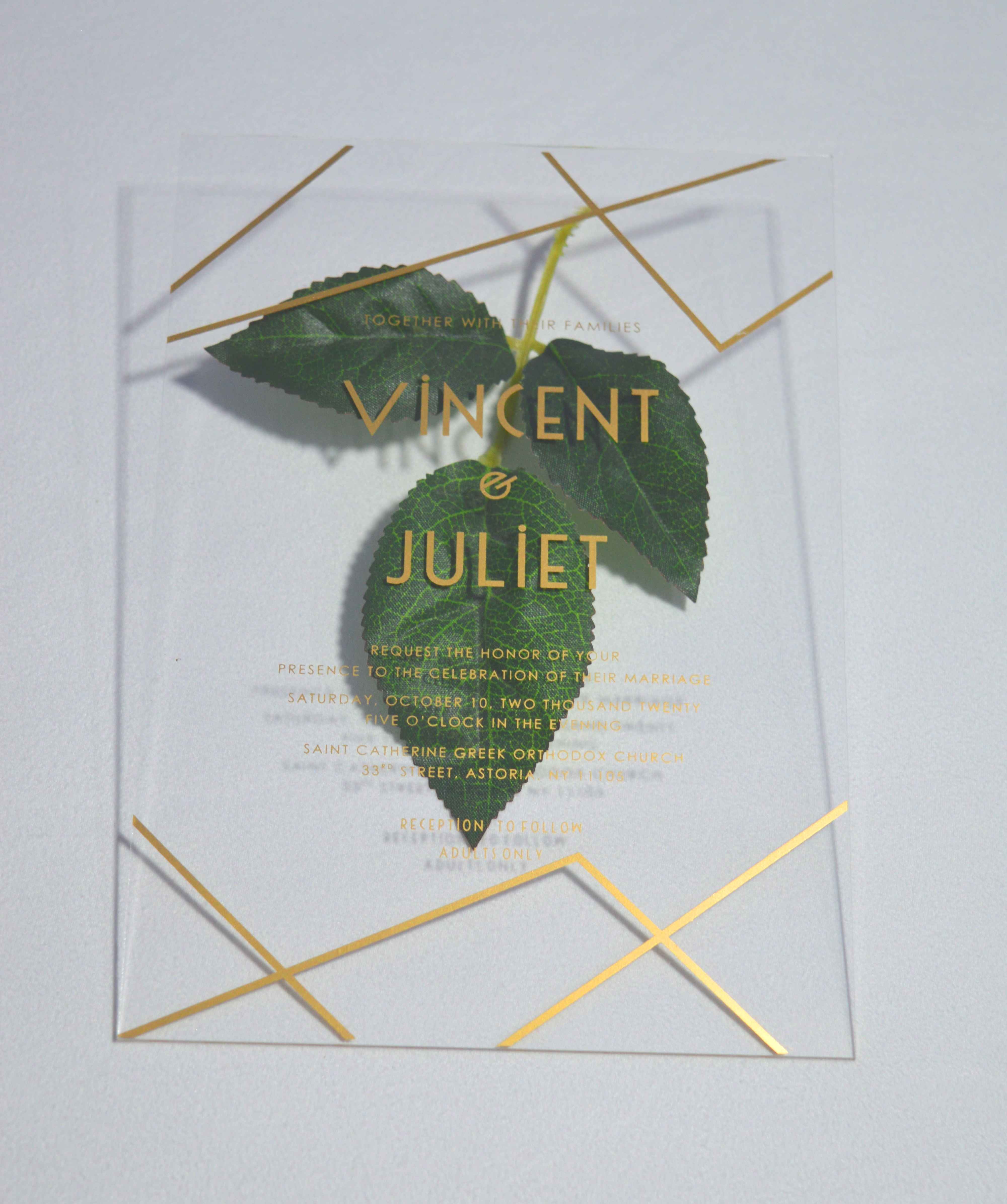 A clear, rectangular acrylic wedding invitation is displayed on a light gray surface, flanked by artificial green leaves. The invitation features elegant gold text and a minimalist gold geometric design consisting of intersecting lines in the top right and bottom left corners.
