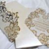 An ornate wedding invitation suite is displayed on a white surface, featuring ivory, pink, and transparent acrylic elements decorated with elaborate gold foil filigree.