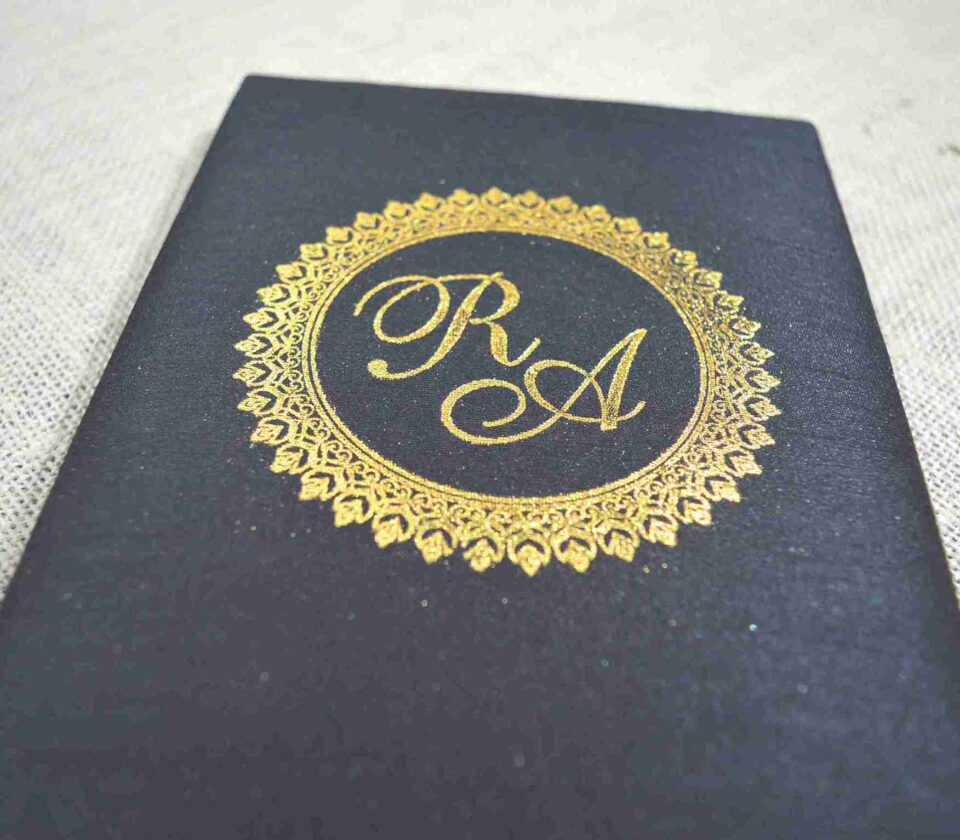 A top-down view of an open, square black silk invitation box. The interior lid features a matching horizontal silk pocket holding a Acrylic card with the names in elegant black script. The base of the box is lined with plush, gathered black fabric. The box is set against a textured white fabric background.