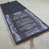 A modern wedding invitation made of clear acrylic, resting on a burlap surface. The invitation features elegant white screen-printed text and is framed by white floral scrollwork borders. A black folio is visible in the blurred background.