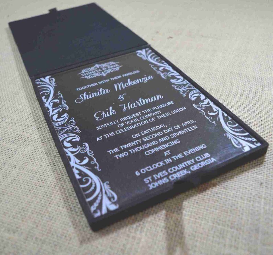 A modern wedding invitation made of clear acrylic, resting on a burlap surface. The invitation features elegant white screen-printed text and is framed by white floral scrollwork borders. A black folio is visible in the blurred background.