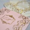 An ornate wedding invitation suite is displayed on a white surface, featuring ivory, pink, and transparent acrylic elements decorated with elaborate gold foil filigree.