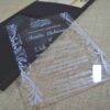 A modern wedding invitation made of clear acrylic, resting on a burlap surface. The invitation features elegant white screen-printed text and is framed by white floral scrollwork borders. A black folio is visible in the blurred background.