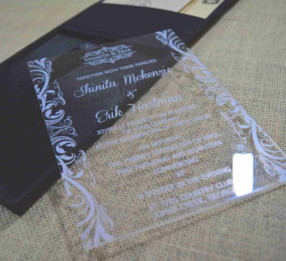 A modern wedding invitation made of clear acrylic, resting on a burlap surface. The invitation features elegant white screen-printed text and is framed by white floral scrollwork borders. A black folio is visible in the blurred background.
