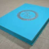 An angled side view of a closed, rectangular turquoise silk invitation box resting on a textured, neutral-toned fabric surface. The flat lid is centered with a shimmering silver foil monogram featuring the initials in an elegant script. The initials are enclosed within a circular, decorative silver border with a delicate, lace-like pattern. The clean lines of the box and the vibrant turquoise color give the invitation a modern yet sophisticated appearance.